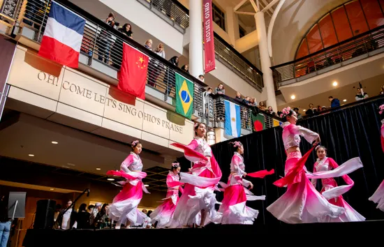 Dancers in pink and white traditional attire perform on stage beneath international flags and an Ohio State banner.